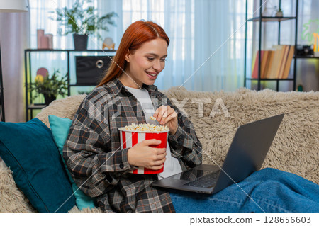 Woman sitting on couch watching movie on laptop eating popcorn laughing emotionally with excitement Woman sitting on couch watching movie on laptop eating popcorn laughing emotionally with excitement 128656603