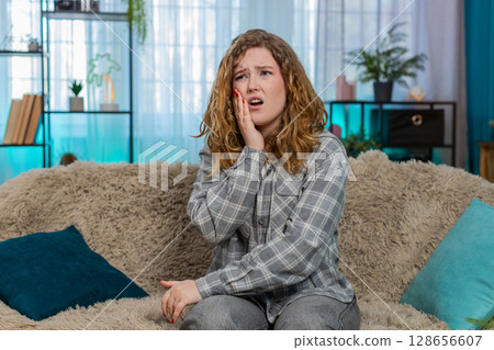 Woman sitting on sofa holding cheek with hand suffering from sharp toothache feeling pain and stress Woman sitting on sofa holding cheek with hand suffering from sharp toothache feeling pain and stress 128656607
