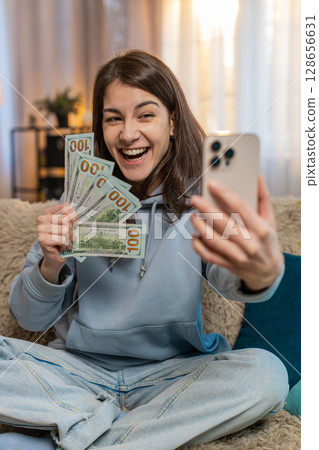 Smiling woman blogger influencer recording story on smartphone sitting on sofa showing money at home 128656631