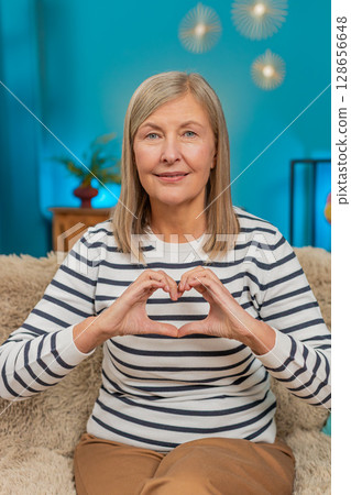 Mature woman making heart gesture on sofa, expressing love and gratitude, showing affection at home 128656648