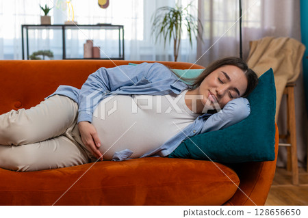 Smiling young pregnant woman stroking big belly while sleeping on comfortable sofa dreaming at home 128656650