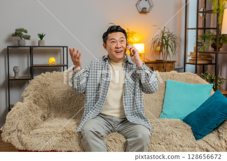 Smiling young Chinese man making fun phone conversation with friends sitting on couch in living room 128656672