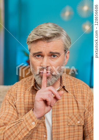 Mature man sitting on sofa making quiet gesture, expressing seriousness and secrecy at home 128656686
