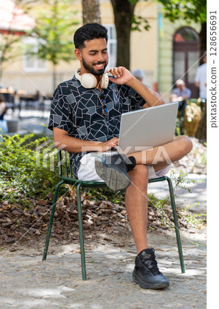 Indian man in headphones working online distant job with laptop browsing website chatting outdoors 128656691