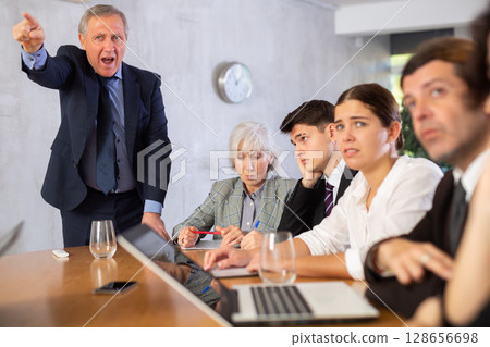 Outraged old manager getting mad at young adult mature office workers sitting with reports and laptops at table in meeting room Outraged old manager getting mad at young adult mature office workers sitting with reports and laptops at table in meeting room 128656698