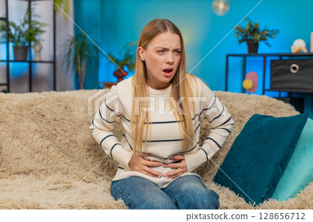 Caucasian woman abdominal stomach pain on sofa holding belly cramps suffering indoors discomfort 128656712