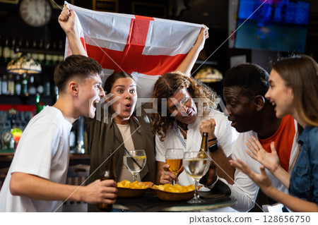 Males and females with England flag clinking glasses with beer on party in bar Males and females with England flag clinking glasses with beer on party in bar 128656750