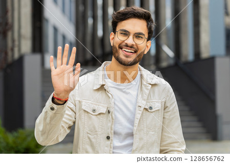 Indian man smiling friendly at camera, waving hands hello, hi, greeting or goodbye in city street Indian man smiling friendly at camera, waving hands hello, hi, greeting or goodbye in city street 128656762