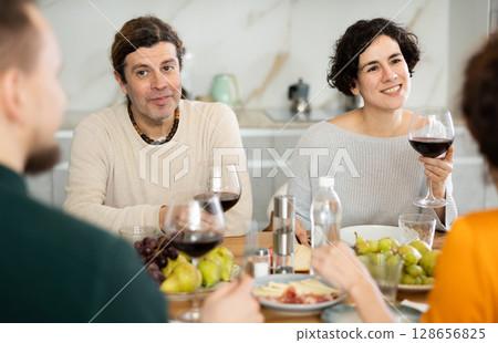 Group of joyful individuals are celebrating a holiday, enjoying drinks and conversation around table in their home 128656825
