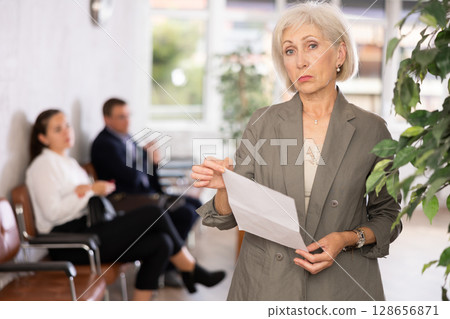 Upset elderly woman with documents standing in reception 128656871