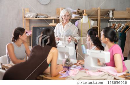 Mature female teacher speaking students during dressmaking class Mature female teacher speaking students during dressmaking class 128656916
