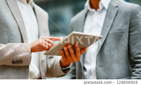 Two men wearing suits. One of the men is holding a tablet and pointing to the screen.	 128656928