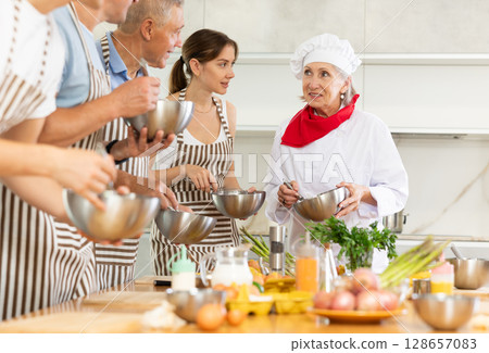 Smiling old woman chef of cooking course teaching attendees how to mix sauce 128657083