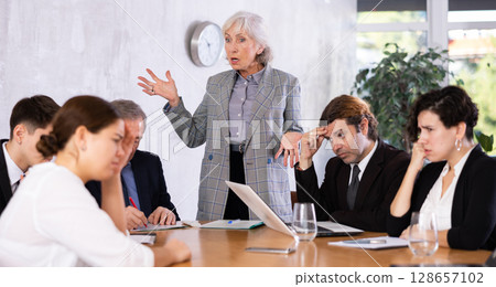 Outraged old female manager getting mad at office workers sitting with reports and laptops at table in meeting room 128657102