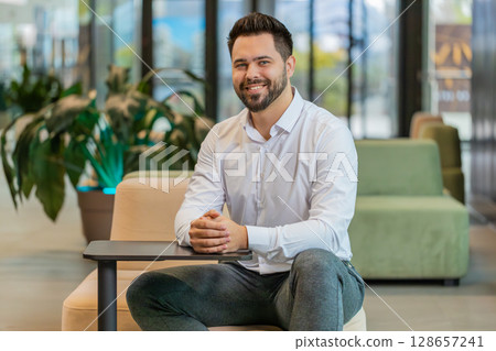 Portrait of smiling young businessman looking at camera sitting in hotel lobby Portrait of smiling young businessman looking at camera sitting in hotel lobby 128657241