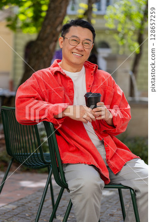 Happy Asian man enjoying drinking morning coffee hot drink, relaxing, taking a break on city street Happy Asian man enjoying drinking morning coffee hot drink, relaxing, taking a break on city street 128657259