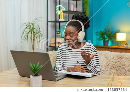 African American woman studying online headphones notes laptop sofa home table indoors education African American woman studying online headphones notes laptop sofa home table indoors education 128657384