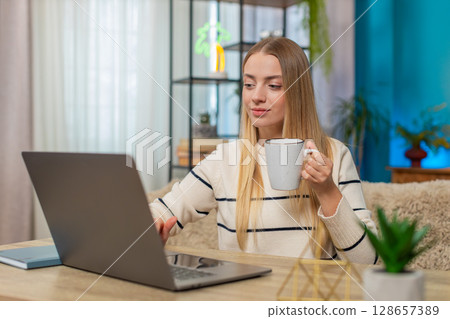 Caucasian woman freelancer working remotely sitting at home table typing on laptop drinking coffee Caucasian woman freelancer working remotely sitting at home table typing on laptop drinking coffee 128657389