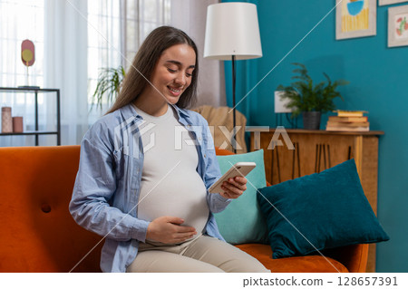 Smiling pregnant woman using smartphone making online purchases for baby sitting on home couch 128657391
