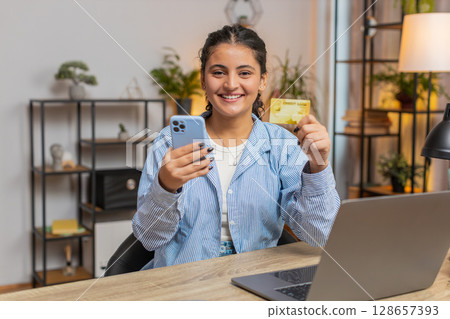 Happy Indian woman freelancer using credit card and smartphone for online shopping at table 128657393