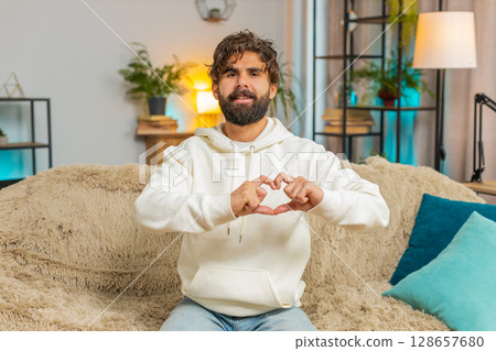 Happy Indian young man at home living room couch makes symbol of love, showing heart sign to camera 128657680