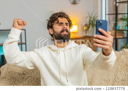 Happy young man using cellphone celebrating success victory winning lottery on sofa in living room 128657778