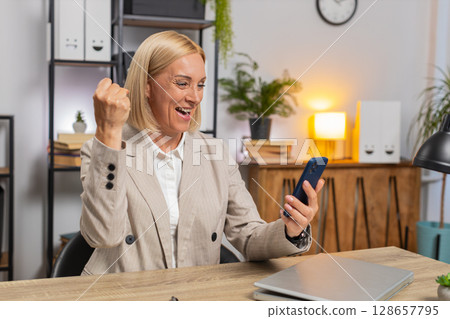 Happy mature businesswoman celebrating success by reading great news on smartphone at home office Happy mature businesswoman celebrating success by reading great news on smartphone at home office 128657795
