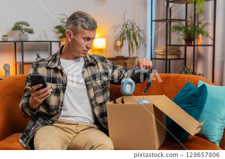 Unhappy man unpacking carton box feeling dissatisfied with getting damaged wireless headphones home Unhappy man unpacking carton box feeling dissatisfied with getting damaged wireless headphones home 128657806