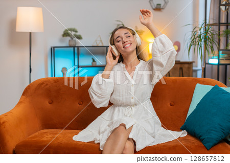 Joyful young Caucasian woman listening to music through wireless headphones sitting on sofa couch Joyful young Caucasian woman listening to music through wireless headphones sitting on sofa couch 128657812