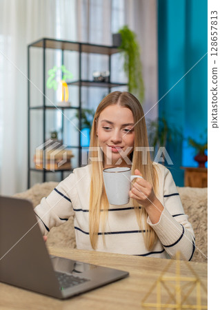 Caucasian woman freelancer working remotely sitting at home table typing on laptop drinking coffee 128657813
