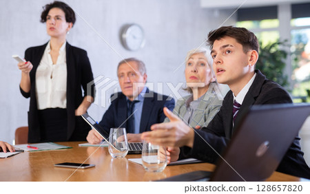 Concentrated young adult old office workers sitting around the table and looking at screen while Latin female using projector remote control in office Concentrated young adult old office workers sitting around the table and looking at screen while Latin female using projector remote control in office 128657820