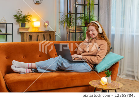 Happy relaxed Caucasian young woman in wireless headphones listening music using laptop on sofa 128657843