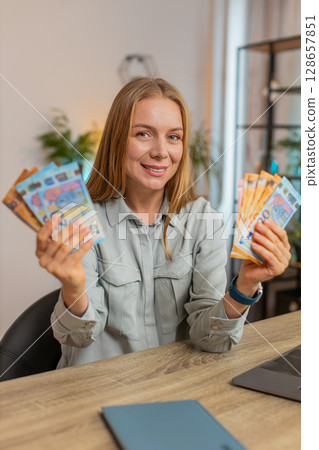 Excited businesswoman freelancer holding fan of euro cash and dancing sitting at home office table 128657851