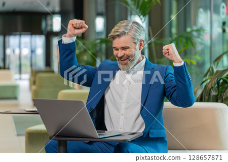 Caucasian bearded businessman working on laptop surprised by sudden victory in modern hotel lobby Caucasian bearded businessman working on laptop surprised by sudden victory in modern hotel lobby 128657871