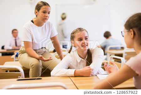 Teenage girls classmates communicating in classroom during break Teenage girls classmates communicating in classroom during break 128657922