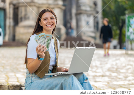 Happy rich woman using laptop winner victory holding cash money in dollar banknotes on city street Happy rich woman using laptop winner victory holding cash money in dollar banknotes on city street 128658777