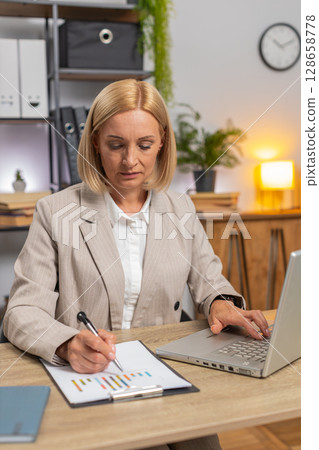 Focused mature businesswoman using laptop working with financial graph documents at home office desk 128658778