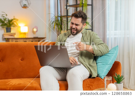 Upset young Caucasian man freelancer using laptop engages in heated conversations sitting on sofa Upset young Caucasian man freelancer using laptop engages in heated conversations sitting on sofa 128658863