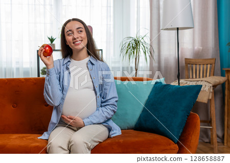Happy pregnant woman eating fresh red apple fruit sitting on sofa healthy vitamin food motherhood 128658887