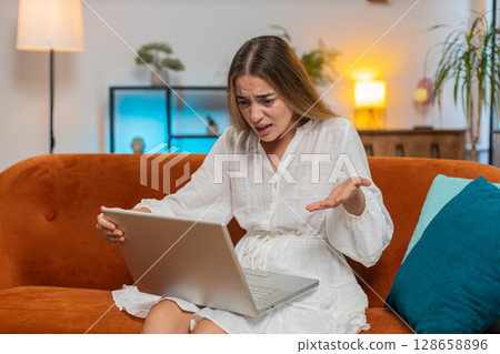 Upset young Caucasian woman using laptop computer having anxiety and stress sitting on sofa at home Upset young Caucasian woman using laptop computer having anxiety and stress sitting on sofa at home 128658896