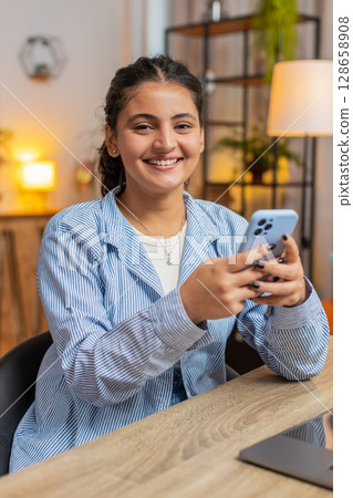 Smiling young woman freelancer sitting at table using smartphone texting in living room at home Smiling young woman freelancer sitting at table using smartphone texting in living room at home 128658908
