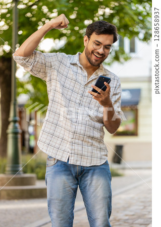 Hispanic young man using smartphone celebrating win good message news clenching fist on city street Hispanic young man using smartphone celebrating win good message news clenching fist on city street 128658937