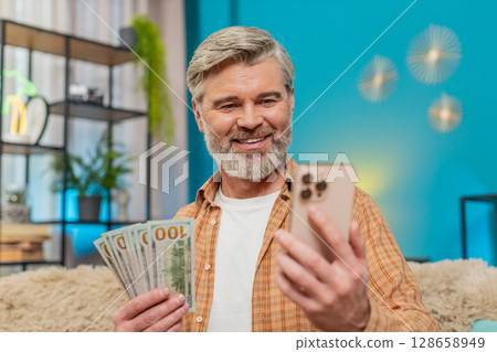 Mature man with smartphone and money, making financial transfer via mobile app, smiling at home Mature man with smartphone and money, making financial transfer via mobile app, smiling at home 128658949