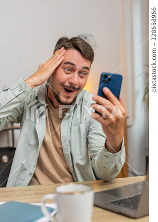 Happy young man freelancer using cellphone celebrating success victory winning lottery at table 128658966