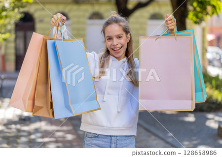 Happy smiling young Caucasian woman shopaholic carrying shopping bags and walking on city street 128658986