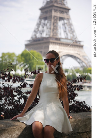Elegant woman in white dress sitting by Seine with Eiffel Tower 128659418
