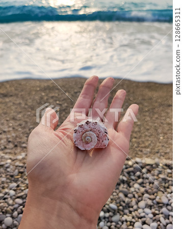 holding a seashell in his hand with a view of the sea and the beach 128665311