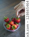 Hands are picking strawberries. Many fresh strawberries in a clear plastic glass lid on a wooden table background, fruit, food, decor, object, copy space 128665464