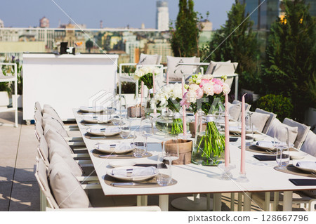 Exquisitely set long dining table on a sunlit rooftop terrace 128667796