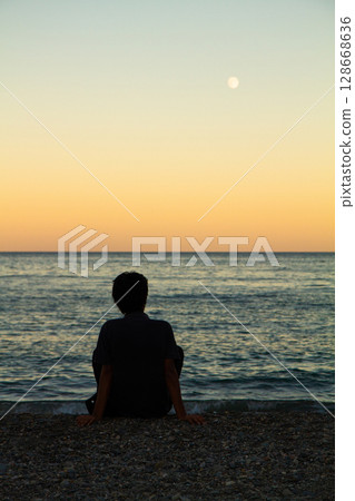 a lonely man sits on the seashore at sunset 128668636